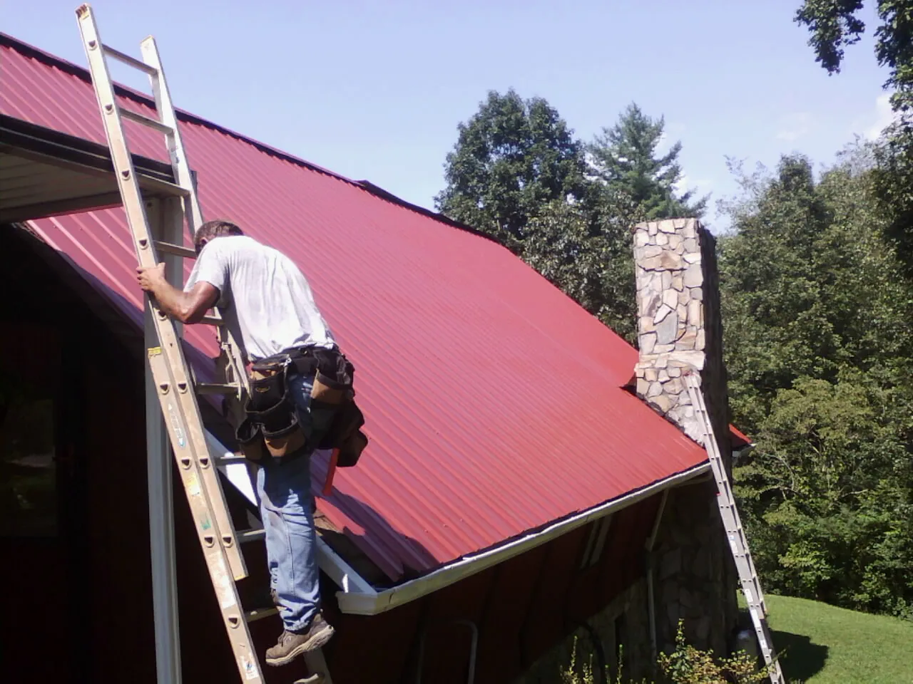 Roofer on a ladder beside a red metal roof and stone chimney — Soffit & Fascia Repair by Roofing Brothers in Convent Station